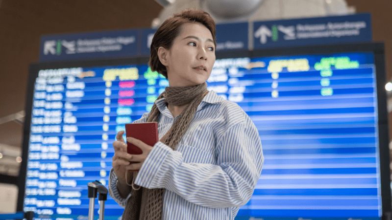 woman standing inside airport in front of the screen, searching for her flight schedule with ticket in her hand.
